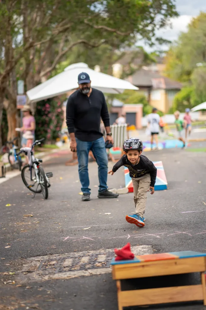 Young boy running toward a cornhole game while adults watch during a local street activity day