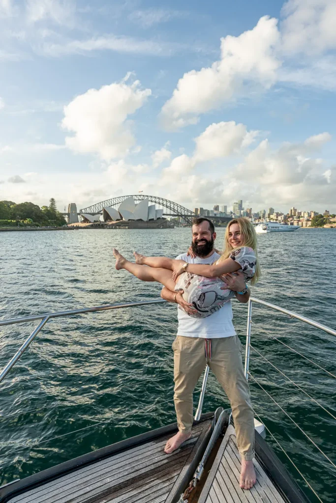 Man holding a woman on the bow of a yacht with Sydney Harbour landmarks behind them