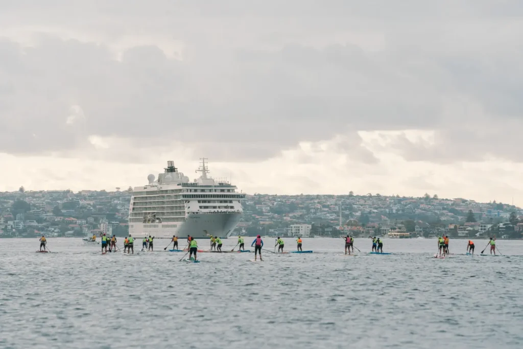 Paddleboard competitors racing across Sydney Harbour with cruise ship backdrop