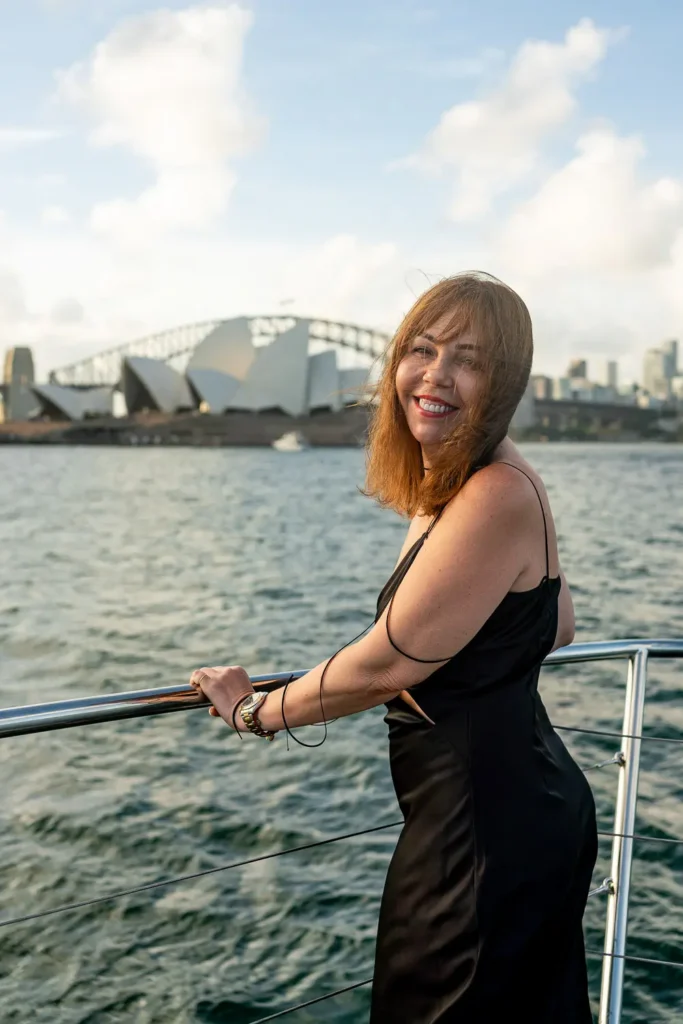 Woman in a black dress smiling on a yacht with the Sydney Opera House and Harbour Bridge behind her