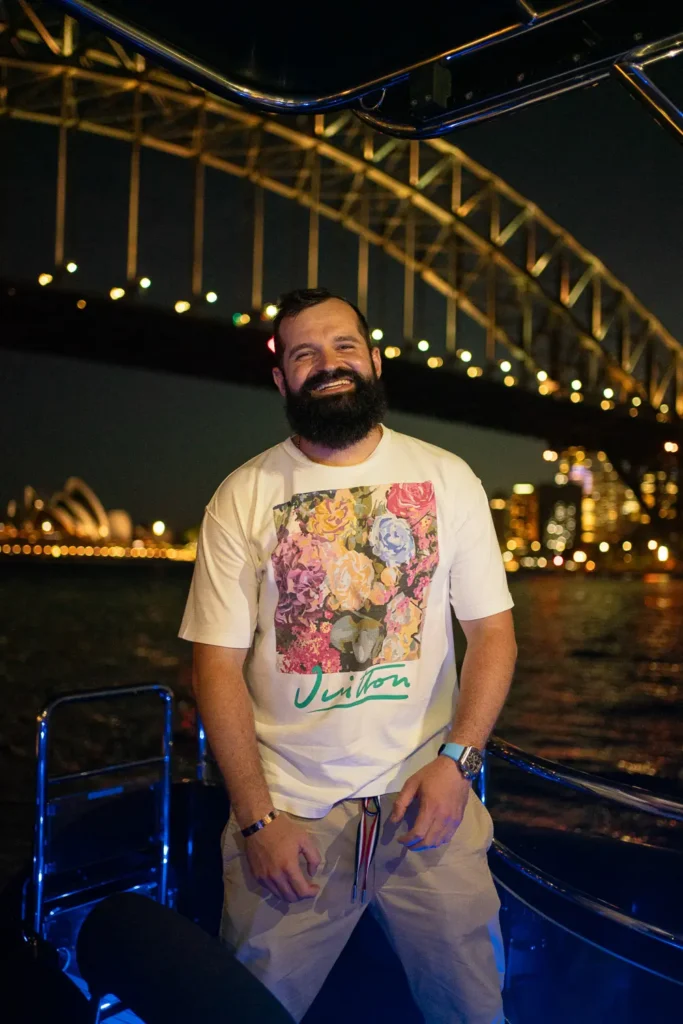 Smiling man posing on a yacht at night with the Harbour Bridge illuminated behind him