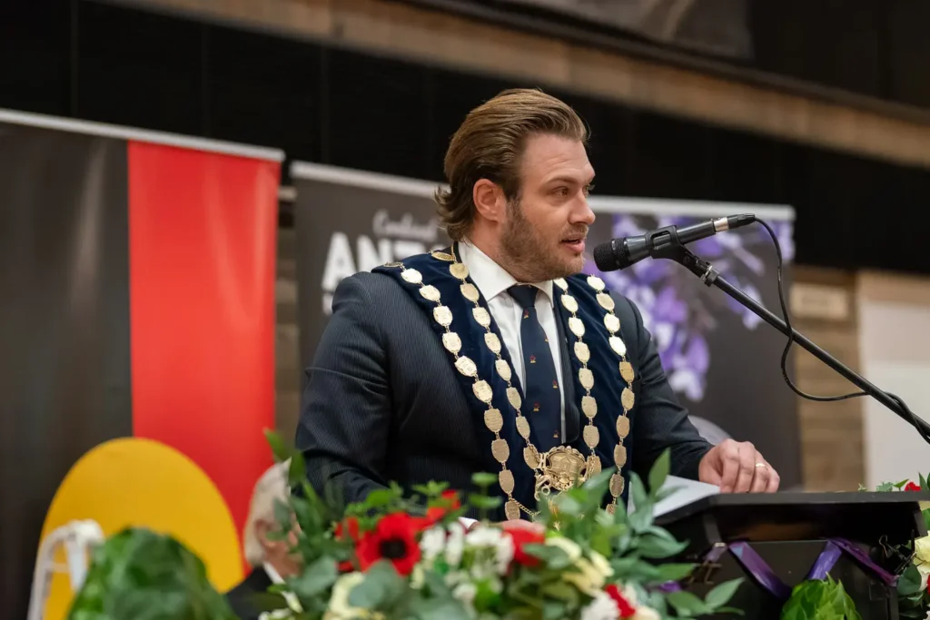 Mayor Zac Miles speaking at podium during Combined ANZAC Memorial Service in Hunters Hill with ceremonial chain and floral arrangements