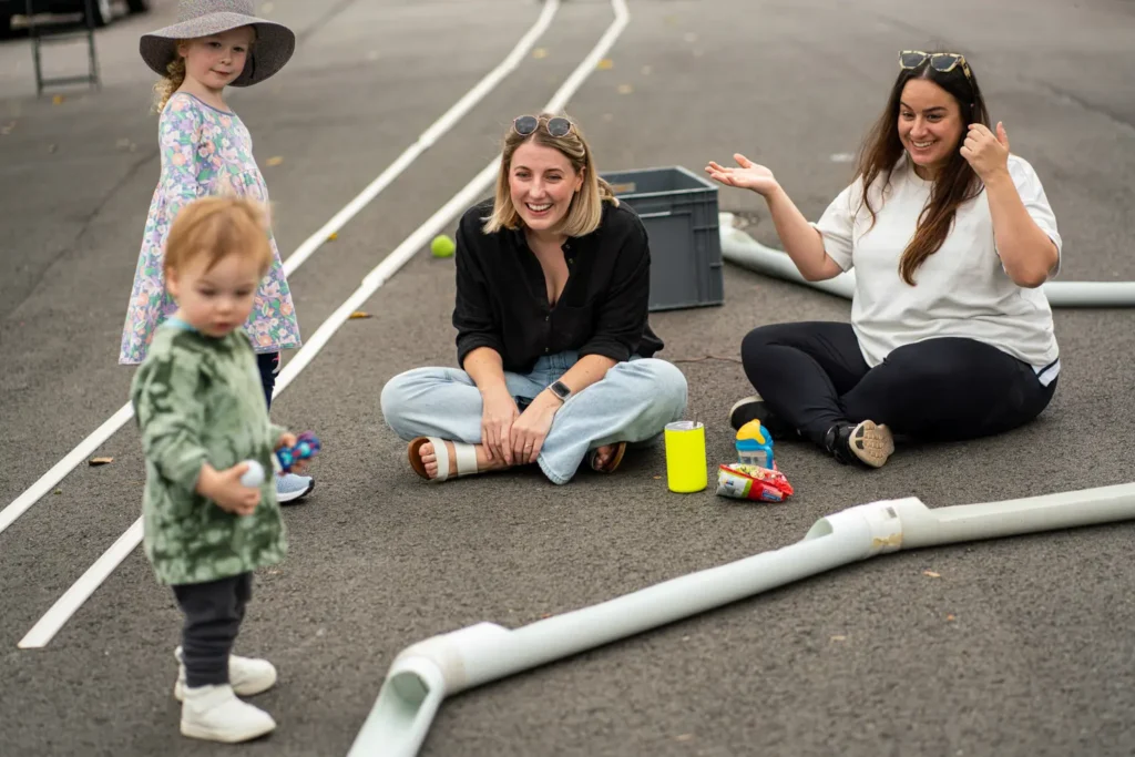 Parents and children sitting on a suburban street beside a temporary toy race track during a community play day
