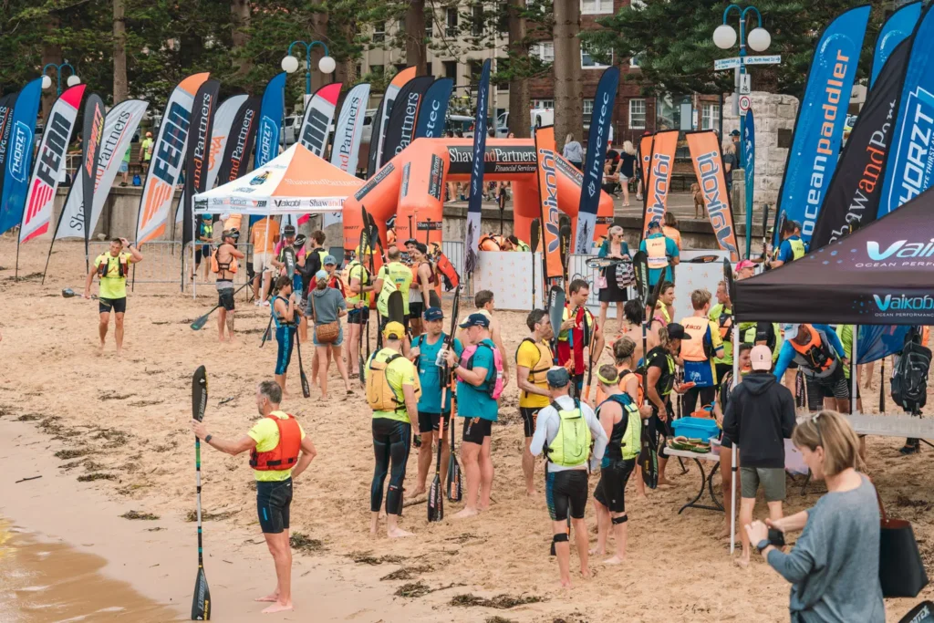 Busy race village setup with surf skis, sponsor flags, and paddlers preparing on the beach