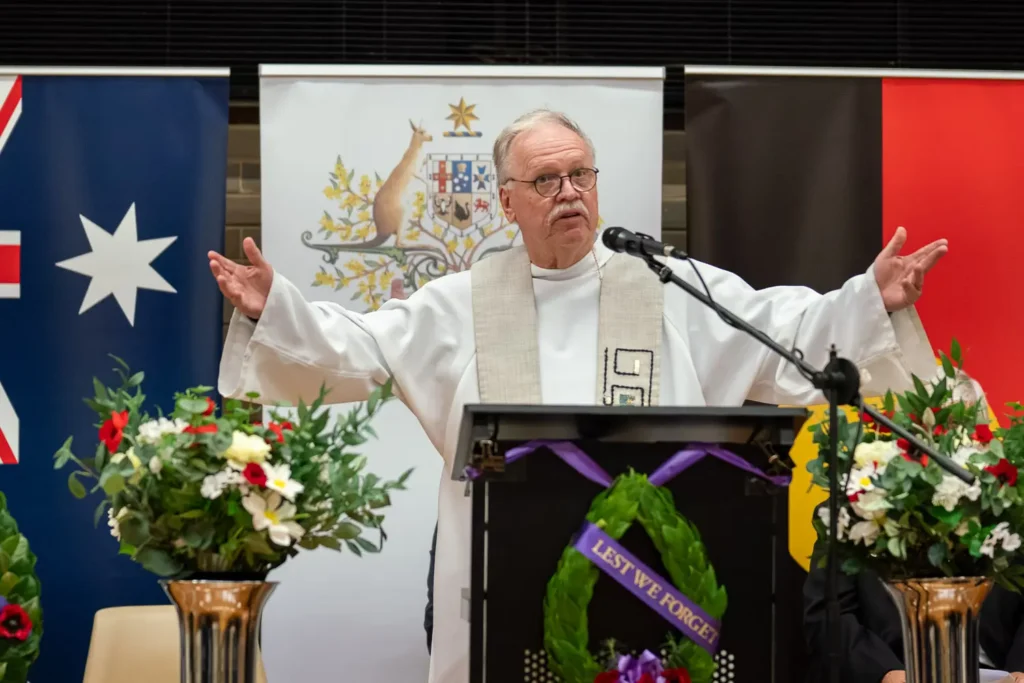 Priest addressing audience at ANZAC memorial service with wreaths and flags in background