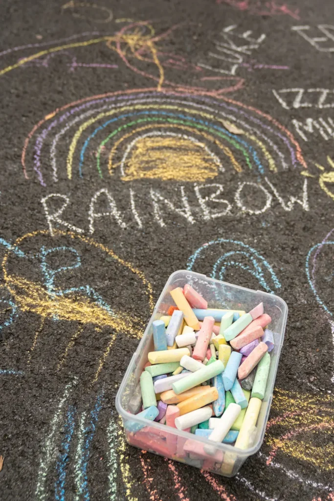 Colourful rainbow chalk drawing on asphalt beside a container of pastel chalk at a children’s street play event