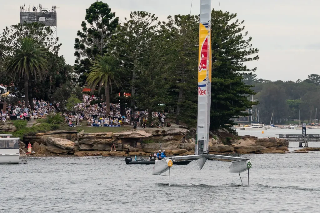 Red Bull Italy SailGP F50 catamaran sailing near Shark Island with spectators watching from the shoreline