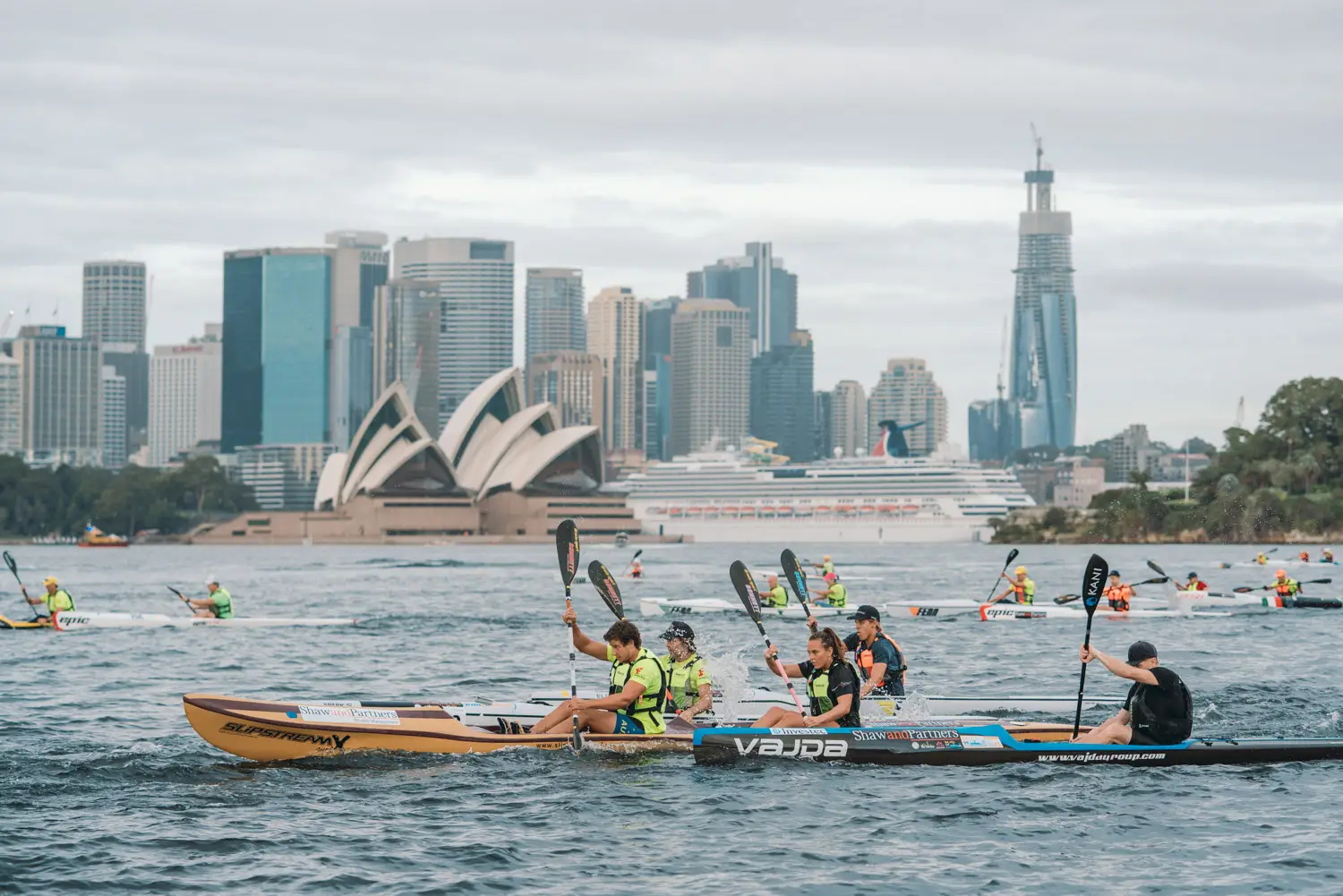 Two surf ski paddlers racing across Sydney Harbour with the Opera House and city skyline behind them