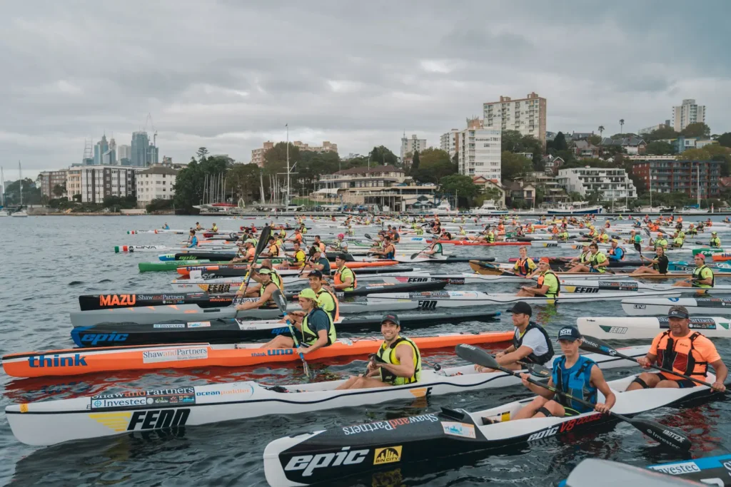 Large group of surfski paddlers gathered at the start line on Sydney Harbour under cloudy skies