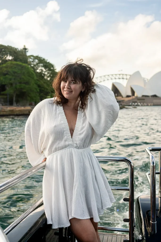 Woman in a flowing white dress posing on a yacht with the Sydney Opera House in the background