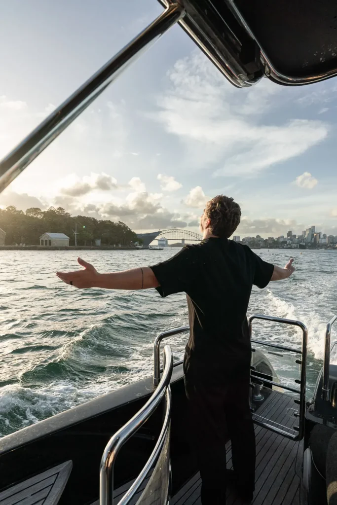 Person standing with open arms on the back of a yacht during a Sydney Harbour sunset cruise