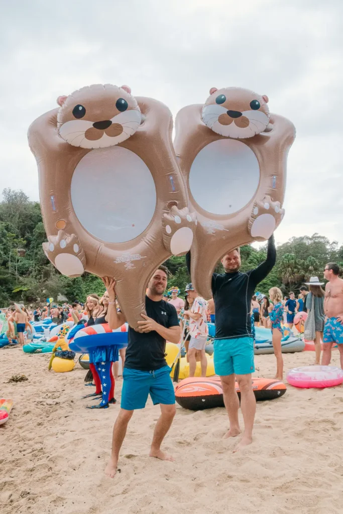 Two participants holding giant otter inflatables on Shelly Beach before the race
