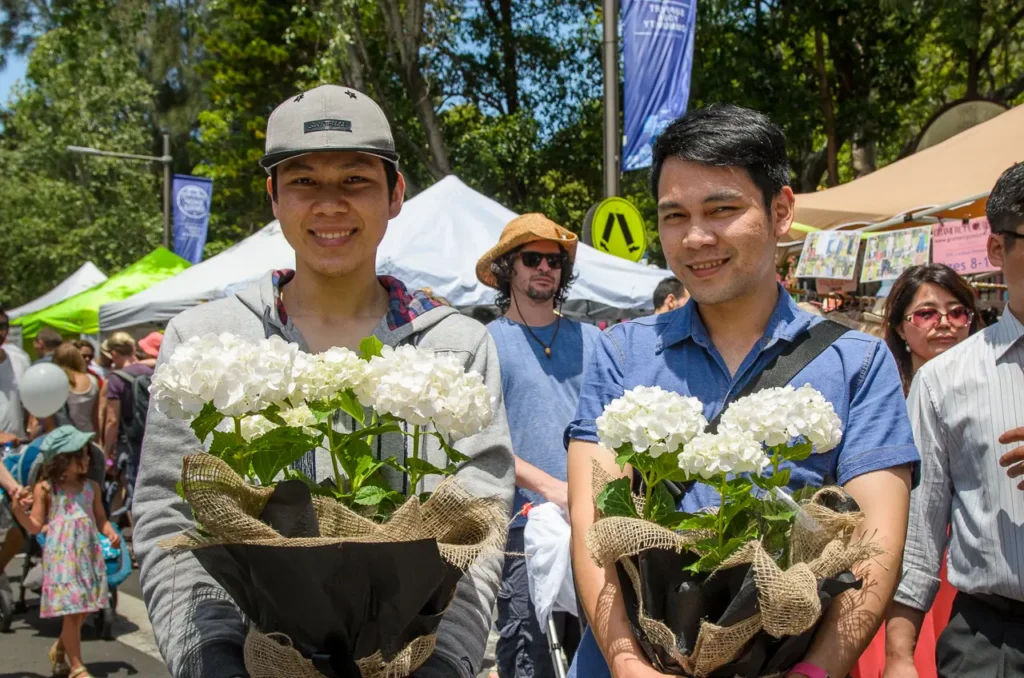 Two smiling visitors holding white hydrangeas among busy market stalls at Glebe Street Fair Sydney