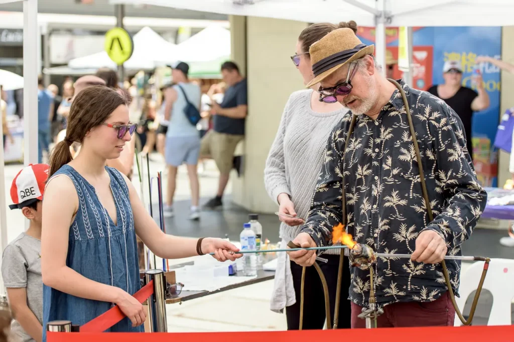 Glass artist demonstrating flame-working techniques to visitors during a creative workshop at Glebe Street Fair
