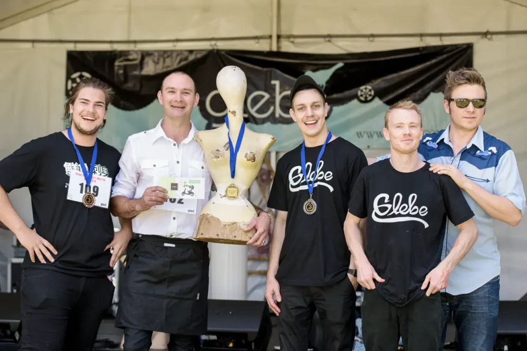 Smiling Greatest Waiter Race competitors celebrating with medals and awards on stage at Glebe Street Fair
